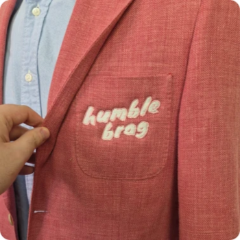 Close-up of a pink blazer pocket embroidered with "humble brag," a hand lifting the flap.